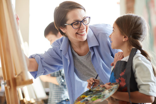Portrait of smiling female teacher helping little girl painting picture on easel in art class, copy space