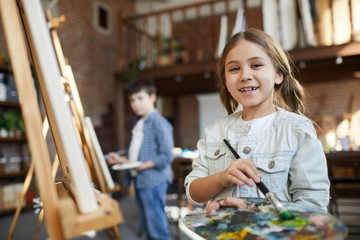 Waist up portrait of cute little girl painting picture on easel in art class and holding palette,...