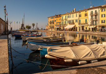 Fototapeta na wymiar Scenic view of the Old Port of the medieval town of Lazise on the shore of Lake Garda with the typical colored houses and moored boats in a sunny day, Veneto, Italy