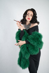 charming graceful pin up girl in a black evening dress and a green fluffy boa in her hands stands and smokes a cigarette with a mouthpiece on a white solid Studio background