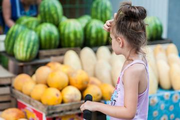 Teenager girl on a scooter stands near the counter with watermelons and melons
