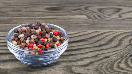 Peppercorns mix. Black, white, green and red pepper corns in a glass bowl on wood background. Piper nigrum. Colorful mixture of dried seasoning close-up in blue transparent dish on brown wooden table.