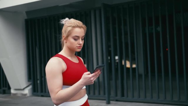 Plus size blond mixed racee smiling woman wearing red sportswear using smartphone outdoors.