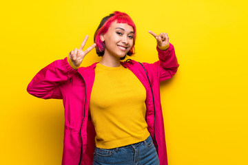 Young woman with pink hair over yellow wall showing victory sign with both hands