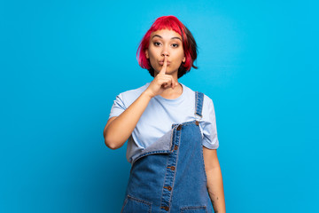 Young woman with pink hair over blue wall showing a sign of silence gesture putting finger in mouth