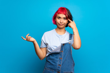 Young woman with pink hair over blue wall making the gesture of madness putting finger on the head