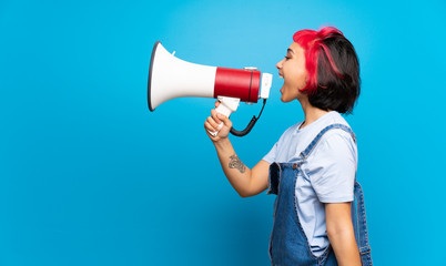 Young woman with pink hair over blue wall shouting through a megaphone