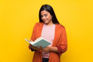 Young Colombian girl over yellow wall holding a book and enjoying reading