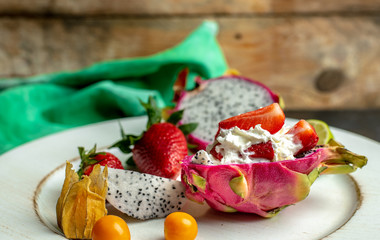 on a white, wooden plate, lie the fresh fruit of strawberries, among them a beautiful, pink dragon fruit filled with whipped cream and strawberries