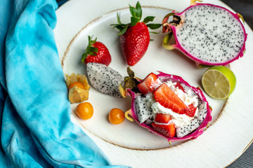 on a white, wooden plate, lie the fresh fruit of strawberries, among them a beautiful, pink dragon fruit filled with whipped cream and strawberries