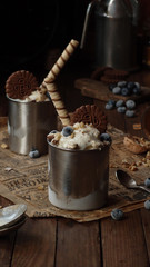 close up of a homemade icecream dessert in a metall jar whith blueberries, chocolate cookies, nuts and wafer roll on a rustic napkin on the wooden table. low key photo