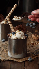 close up of a homemade icecream dessert in a metall jar whith blueberries, chocolate cookies, nuts and wafer roll on a rustic napkin on the wooden table. low key photo