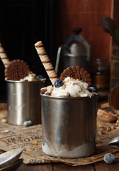 close up of a homemade icecream dessert in a metall jar whith blueberries, chocolate cookies, nuts and wafer roll on a rustic napkin on the wooden table. low key photo