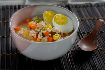 Hot boiled rice soup with egg, corns, green peas and carrots in bowl on wooden table. Delicious Thai food for served in breakfast. 