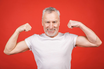 Check this out! Senior man showing muscle. Aged man flexing his arms isolated against red background. Cheerful excited modern cool pensioner grandpa practising bodybuilding.