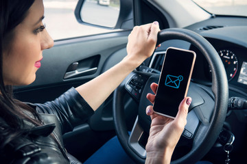 Closeup portrait of young woman driving in car and checking her email on phone. Girl holding wheel...
