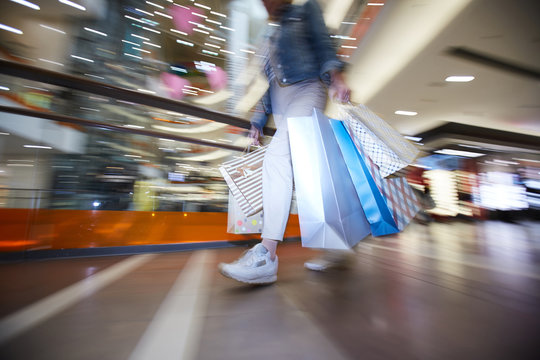 Below view of unrecognizable hipster girl in sneakers moving over shopping mall and carrying colorful paper bags with clothing bought on seasonal sale