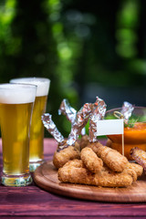 Chicken and Cheese Set for beer and two glasses of beer. Chicken wings, cheese sticks and cheese balls with bright vegetable sauce. On the cutting board, wooden and green summer background