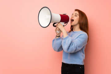 Young redhead woman over pink background shouting through a megaphone
