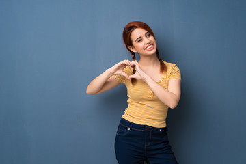 Young redhead woman over blue background making heart symbol by hands