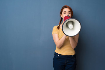 Young redhead woman over blue background shouting through a megaphone