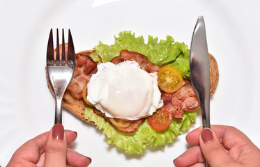 Women's hands hold a knife and fork against the background of a beautiful sandwich with fried bacon, egg and cherry tomatoes. Unhealthy food for breakfast. CLose-up