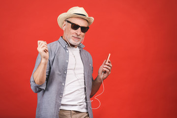 Portrait of senior happy cheerful elderly man dancing and listening music witn phone and headphones  isolated against red background.