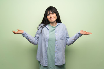 Young woman over green wall having doubts while raising hands and shoulders