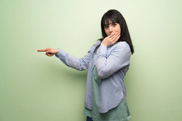 Young woman over green wall pointing finger to the side with a surprised face