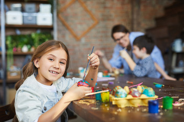 Portrait of smiling girl looking at camera while painting eggs for Easter in art studio, copy space