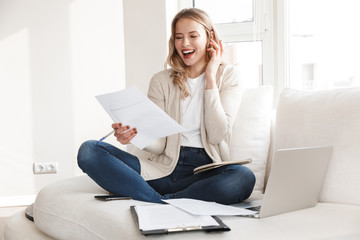 Beautiful blonde woman posing sitting indoors at home using laptop computer talking by mobile phone.