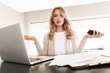 Confused blonde woman posing sitting indoors at home using laptop computer holding mobile phone.