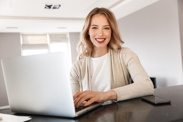 Beautiful blonde woman posing sitting indoors at home using laptop computer.