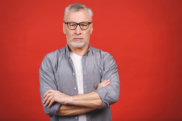 Portrait of a mature serious businessman with crossed hands wearing glasses isolated against red background. Happy senior man looking at camera with copy space. 