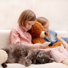 Two little girls (sisters 7 and 8 years old) are watching an e-book and a smartphone at home on the sofa with their cat.