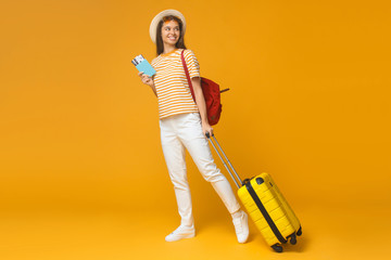 Young female tourist standing with suitcase and backpack, isolated on yellow background. Travel concept