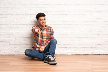 Young man sitting on the floor points finger at you with a confident expression