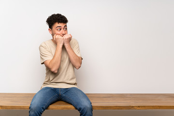 Young man sitting on table nervous and scared putting hands to mouth