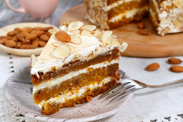 Close-up the slice  of vegetarian carrot cake with almonds on a saucer and a fork near. Pink cup, plate with almonds and wooden board with cake in the background.