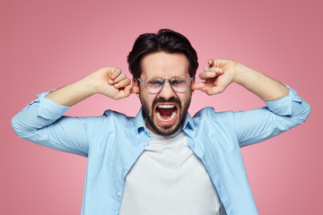 Irritated handsome male plugs ears and screaming to ignore a conflict, expresses negativeness, opens mouth, closes eyes, ignores annoying sound, stands over pink background. Negative emotions.