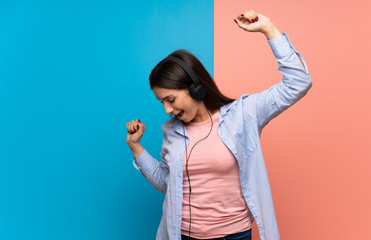 Young woman over pink and blue wall listening to music with headphones and dancing