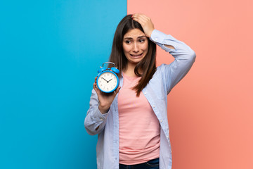 Young woman over pink and blue wall restless because it has become late and holding vintage alarm...