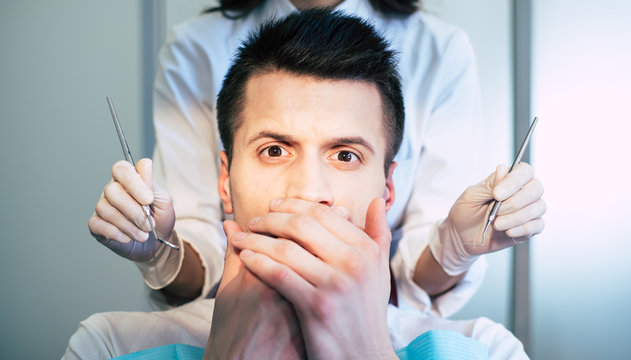 Nixie! A man who is scared of dental treatment  is sitting in a dental chair and covering his mouth with hand and looking scurrying into the camera.