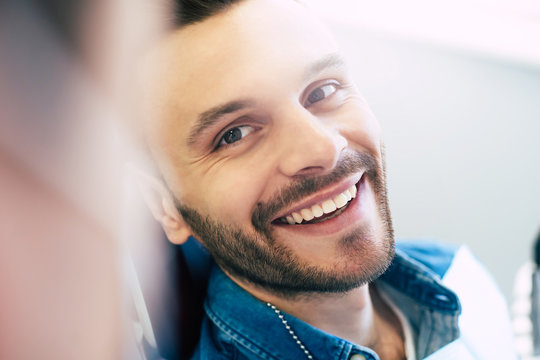 Owner Of A Smile. Hilariously Handsome Man Clothed In A Denim Shirt Is Sitting In A Dental Hair Showing To The Doctor His New Eye Catching Healthy Smile.