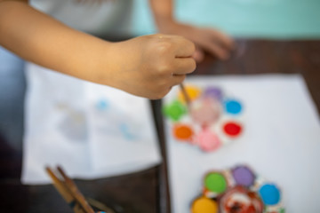 Child's hand put paint brush into used watercolor in palette on desk with background blur