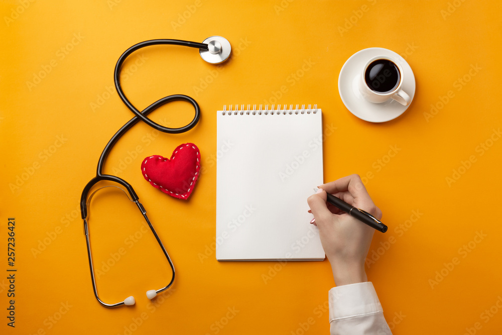 Wall mural Professional doctor writing medical records in a notebook with stethoscope, coffee cup, syringe and heart