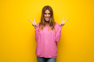 Woman with pink sweater over yellow wall smiling and showing victory sign with both hands