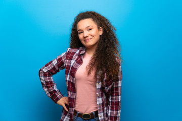 Teenager girl over blue wall posing with arms at hip and smiling