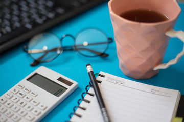 Closeup of blue office desk, keyboard and calculator, cup of tea and notepad.