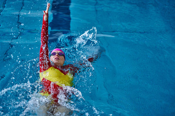 Female athlete in a red-yellow swimsuit is swimming on his back. Splashes of water scatter in different directions.
